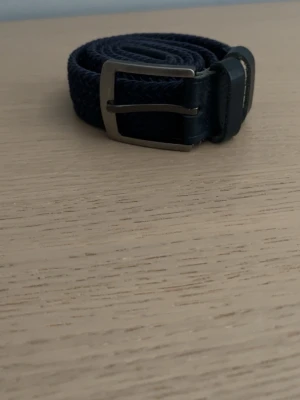 Mörkblått flätat bälte med metallspänne - Snyggt mörkblått flätat bälte med silverfärgat metallspänne och detaljer i skinn. Perfekt för att ge din outfit en clean och stilren touch. Passar till jeans, chinos eller shorts.