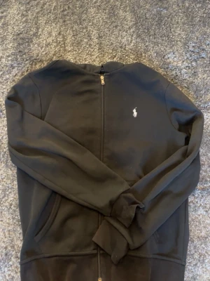 Svart zip hoodie från Polo Ralph Lauren - Snygg svart hoodie med dragkedja från Polo Ralph Lauren. Klassisk design med vit broderad logga på bröstet, två fickor framtill och huva med dragsko. Perfekt för en chill och stilren look. Tillverkad i mjukt bomullsmaterial. 170/92A