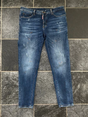Blåa Dsqaured2 jeans - Säljer ett par blåa Dsqaured2 jeans med coola slitningar och färgstänk i orange och vitt. Jeansen har klassisk femficksmodell, normal midja och raka ben. Materialet är denim i bomull och passformen är relaxed fit. Perfekta för en avslappnad streetstyle-look. Storleksguide finns på nätet.