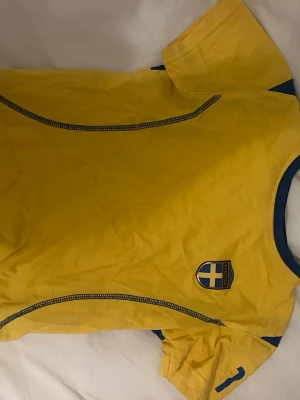 Gul Sverige fotbollströja - Säljer en gul fotbollströja med blå detaljer och Sverige-emblem på bröstet. Perfekt för att heja fram landslaget eller till träningen. !!Om du köper välj gärna att hämta upp på upphämtningsställe istället för hemleverans då det blir enklare för mig att posta på det viset!!