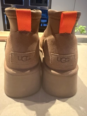 Bruna UGG boots med platåsula - Säljer ett par bruna boots från UGG med kraftig platåsula och mockaöverdel. Skorna har en tydlig orange dragflik baktill och UGG-logga på hälen. Yttersulan är mönstrad för extra grepp. Perfekta för dig som gillar en chunky stil.