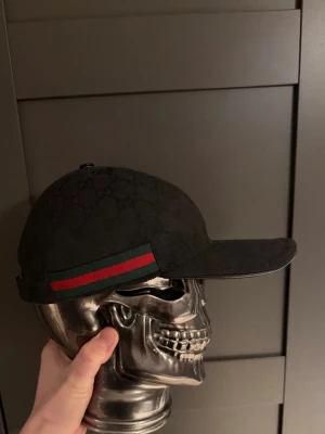 Svart Gucci keps. - Snygg svart keps från Gucci med diskret GG-logomönster över hela kepsen. På sidan finns en klassisk grön och röd rand. Kepsen har böjd skärm och justerbar passform. Perfekt för dig som vill ha en stilren accessoar med lyxig känsla.(ÄKTA) träffas oxå i Sthlm 
