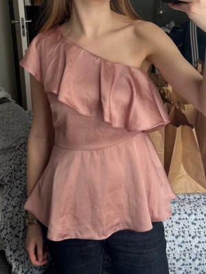 Rosa oneshoulder topp med volang - Snygg rosa oneshoulder topp med bred volangdetalj över axeln och en lätt utsvängd passform. Från hm 
