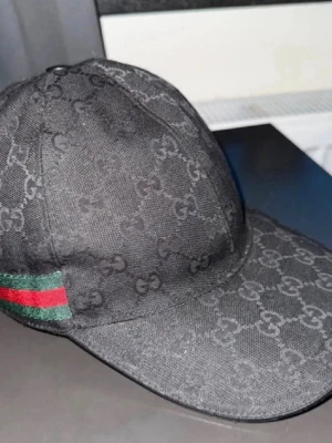 Svart keps från Gucci med monogram - Svart keps från Gucci med klassiskt GG-monogram över hela kepsen och en grön-röd rand på sidan. Kepsen har böjd skärm och är tillverkad i ett slitstarkt tygmaterial. Perfekt för dig som vill ha en lyxig och trendig accessoar.