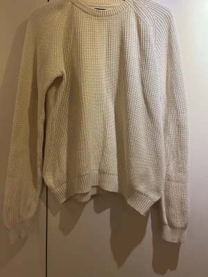 Vit stickad tröja från Lyle & Scott - Säljer en stilren vit stickad tröja från Lyle & Scott med ribbade muddar och rund hals. Tröjan har ett diskret svart tygmärke med gul logga på ärmen och är gjord i ett mjukt, tjockt material som passar perfekt till kyligare dagar.