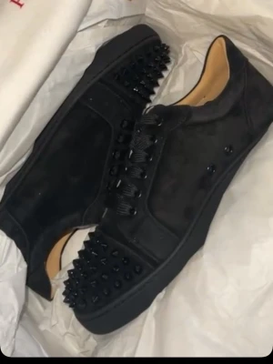 Svarta spikes-sneakers Louboutin - Säljer ett par svarta sneakers från Christian Louboutin med mattsvarta nitar över tån och sidorna. Skorna har en låg siluett, svarta skosnören och en lyxig känsla med läderdetaljer på insidan. Perfekta för dig som vill sticka ut med edgy stil.