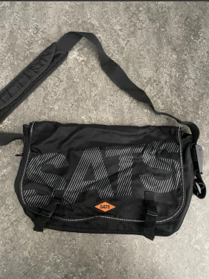 Svart axelväska från SATS - Cool svart axelväska från SATS med stort logotryck på framsidan. Väskan har justerbar axelrem med vaddering, praktiskt spänne och flera fack. Tillverkad i slitstark textil och perfekt för gymmet eller vardagen.