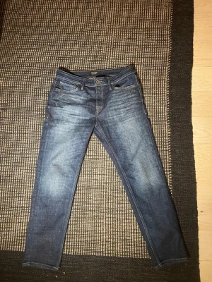 Jack and Jones  - Säljer ett par klassiska blå jeans från Nudie Jeans med snyggt slitna detaljer och femficksdesign. Jeansen har raka ben och är tillverkade i denim med en cool tvätt. Perfekt för dig som gillar en avslappnad och stilren look.