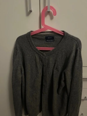 Grå kofta i lammull från GANT - Mysig grå kofta från GANT i superfin lammull. Klassisk rund halsringning och knäppning framtill med ribbade muddar. Perfekt för lager-på-lager och enkel att matcha med olika outfits.