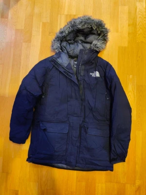 Jacka från The North Face - Jacka från The North Face med huva och fuskpäls. Jackan har lång ärm och klassisk parkasdesign, perfekt för kalla dagar. Logga på ryggen och insidan. Herrjacka. Inga byten.