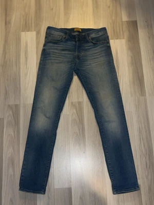 Blå jeans från Jack & Jones - Säljer ett par klassiska blå jeans från jack & jones med fem fickor och normal passform. Jeansen har en snygg tvätt med lätt slitning på låren och knäna samt en brun läderpatch bak i midjan. Perfekta till vardagslooken!
