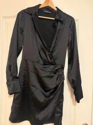 Svart omlottklänning från Stradivarius - Snygg svart omlottklänning från Stradivarius i glansigt satintyg. Klänningen har lång ärm, krage, kort och djup v-ringning. Perfekt för dig som gillar en elegant och trendig look.