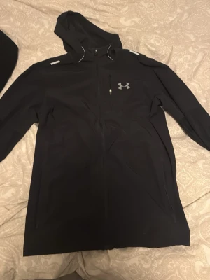 Svart hoodie från Under Armour - Snygg svart hoodie från Under Armour med dragkedja framtill och en liten bröstficka med dragkedja. Loggan i silver på bröstet och reflekterande detaljer på axlarna. Tillverkad i ett mjukt och stretchigt material, perfekt för träning eller chill.