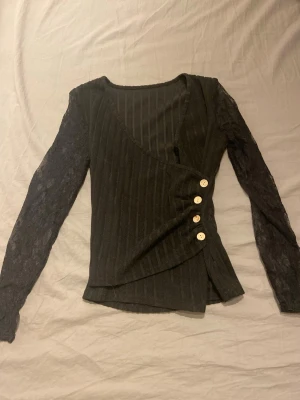 Svart omlottopp med spetsärmar - Snygg svart omlottopp med ribbat tyg och transparenta spetsärmar. Toppen har fyra dekorativa knappar på sidan och en v-ringad halsringning. Perfekt för dig som vill ha en unik och trendig look. Använd ca 2ggr