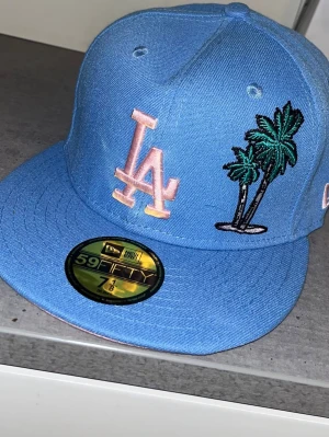 Blå LA Dodgers keps från New Era - Snygg blå 59FIFTY keps från New Era med LA Dodgers-logga i ljusrosa framtill och broderade palmer. Kepsen har platt skärm och broderade detaljer på sidorna, inklusive Los Angeles-märke och MLB-logga. Perfekt för dig som gillar streetwear och sportig stil.
