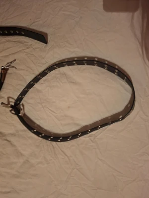 Svart nitar skärp med silverdetaljer - Säljer ett svart skärp med dubbla rader silverfärgade nitar och metallspänne. Skärpet har en edgy vibe och passar perfekt till jeans eller kjol för att ge outfiten lite extra attityd. Materialet är syntet och designen är klassiskt rak.