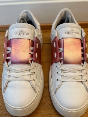 Valentino Garavani vita sneakers - Snygga vita sneakers från Valentino Garavani med metallicrosa detalj över snörningen och på sidorna. Skorna har klassisk låg profil, vita snören och coola nitar bak på hälen. Tillverkade i läder för en lyxig känsla och stilren look.