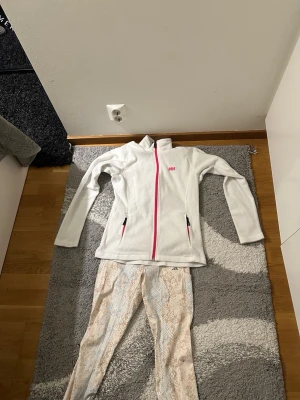Vit fleecejacka Helly Hansen & tights Adidas - Säljer en vit fleecejacka från Helly Hansen med rosa dragkedja och fickor samt ett par Adidas tights med beige och blått mönster. Perfekt för träning eller chill dagar. Jackan har hög krage och tightsen är stretchiga med snyggt print. Helly Hansen koftan är storlek L och byxorna är M.