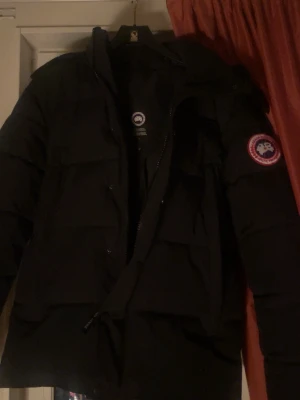 Svart dunjacka från Canada Goose - Säljer en svart dunjacka från Canada Goose med klassisk patch på ärmen. Jackan har huva, dragkedja och knappar framtill samt en puffig, quiltad design som håller dig varm under kalla dagar. Perfekt för dig som vill ha både stil och funktion.