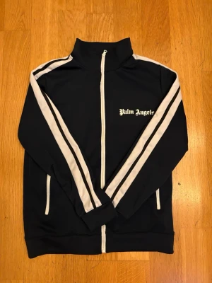 Palm Angels mörkblå track jacket - Mörkblå track jacket från Palm Angels med vita ränder längs ärmarna och vit dragkedja. Hög krage och logga på bröstet i vitt. Sportig och clean design, perfekt till streetwear-stilen. Tillverkad i ett mjukt och stretchigt material.
