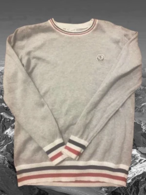 Grå stickad tröja från Moncler - Snygg grå stickad tröja från Moncler med rund halsringning och ribbade muddar. Tröjan har vita, röda och marinblå ränder vid hals, ärmslut och nederkant samt Moncler-logga på bröstet. Perfekt för dig som gillar klassisk stil.