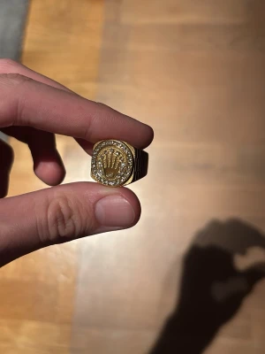 Guldfärgad ring med stenar och logga - Snygg guldfärgad ring med en stor, rund front prydd med gnistrande stenar runt en upphöjd logga. Ringen har en chunky design och känns riktigt lyxig och statement. Perfekt för dig som vill sticka ut och addera lite bling till din stil.