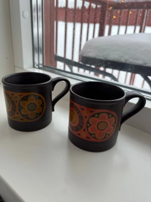 Två retro kaffekoppar med blommönster - Säljer två bruna retro kaffekoppar i keramik med färgglada blommönster i orange, gult och grönt. Kopparna har ett klassiskt handtag och en cylinderformad design, perfekta för dig som gillar vintage och unika detaljer i köket.