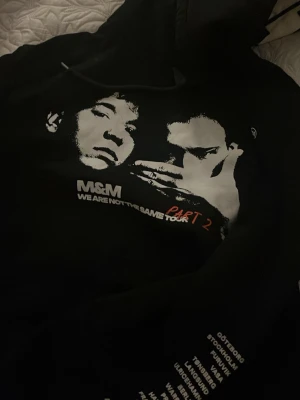 Svart hoodie M&M Tour Part 2 - Svart hoodie med tryck av två ansikten och texten 'M&M WE ARE NOT THE SAME TOUR PART 2' på bröstet. På ena ärmen finns en lista med städer tryckt i vitt. Hoodien har huva och en avslappnad passform, perfekt för en streetwear-look.