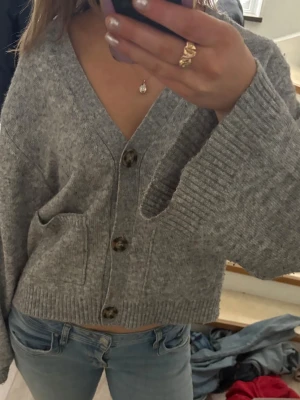 Grå stickad kofta med knappar - Mysig grå stickad kofta med stora bruna knappar och två fickor framtill. Koftan har en croppad passform, v-ringning och ribbade muddar. Perfekt att slänga över en t-shirt eller linne för en chill look. Passar dig som bär S också 