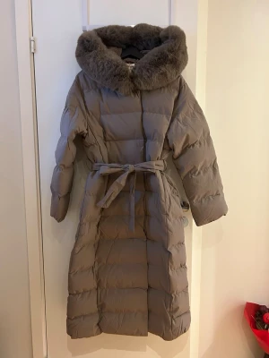 Lång vinterjacka med fuskpäls - Lång vinterjacka med bälte i midjan och stor, fluffig huva kantad med mjuk fuskpäls. Jackan har quiltad design och ger en riktigt varm och stylish look för kalla dagar. Perfekt för dig som vill ha både komfort och trendig vinterstil. 120 cm lång, polyesterfyllning. Stängs med knappar. Ny med prislapp. Nypris runt 2500kr. Mullvadsfärgad 