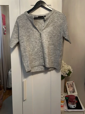 Grå kortärmad kofta från Gina Tricot - Mysig grå kofta i mjukt stickat material från Gina Tricot. Kortärmad modell med rund halsringning och pärlknappar framtill. Perfekt att slänga över en topp för en chill look.