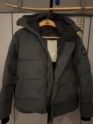 Svart pufferjacka från Canada Goose - Säljer en svart pufferjacka från Canada Goose med hög krage, huva och diskret logotyp på ärmen. Jackan har dragkedja och knappar framtill samt två stora fickor. Perfekt för kalla vinterdagar och riktigt snygg streetstil.