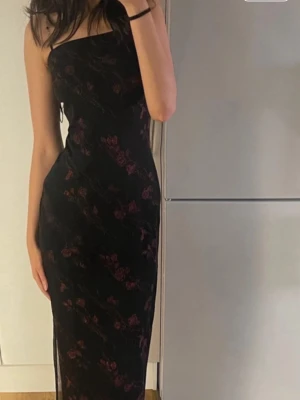 Svart långklänning med blommönster - Elegant svart långklänning med röda blommor och oneshoulder-design. Klänningen har en figurnära passform och är tillverkad i ett tunt, lätt material som ger en snygg siluett. Perfekt för dig som vill sticka ut med en stilren och trendig look.