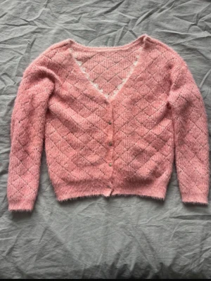 Rosa fluffig kofta med spetsdetalj - Superfin rosa kofta med fluffig känsla och rutmönster. Djup V-ringning med söt vit spetskant och små knappar framtill. Perfekt för lager-på-lager och ger en gullig touch till din outfit.