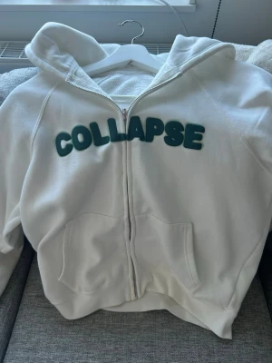 Vit hoodie med grön text  - Vit hoodie med dragkedja med grön text på bröstet. Hoodien har två fickor framtill och en klassisk huva. Tillverkad i mjukt sweatshirtmaterial, perfekt för en avslappnad stil. Hoodien har en liten liten fläck vid dragkedjan men syns knappt. Går säkert och ta bort med vanish eller något annat fläckborttagningsmedel. Storleken är xxs men skulle säga att den passar upp till S då den är stor i storleken