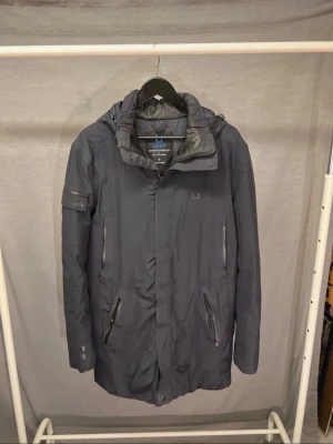 Mörkblå parkas från Ubr - Snygg mörkblå parkas från Ubr med avtagbar huva och flera praktiska fickor med dragkedja. Jackan har en stilren design, hög krage och justerbara detaljer. Perfekt för kalla dagar och tillverkad i ett vattenavvisande material.