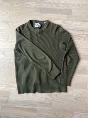 Olivgrön ribbstickad tröja - Snygg olivgrön ribbstickad tröja med rund hals och långa ärmar. Tröjan har en enkel och clean design utan synliga loggor eller detaljer, vilket gör den lätt att matcha med olika outfits. Materialet ser mjukt och stretchigt ut för extra komfort.