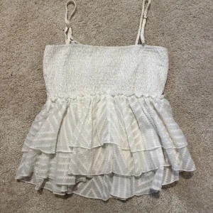 Vit volangtop från SHEIN - Säljer en söt vit linnetopp från SHEIN med tunna axelband och flera lager volanger nedtill. Toppen har ett diskret mönster och smockad överdel som ger en bekväm passform. Perfekt för dig som gillar en feminin och luftig stil.