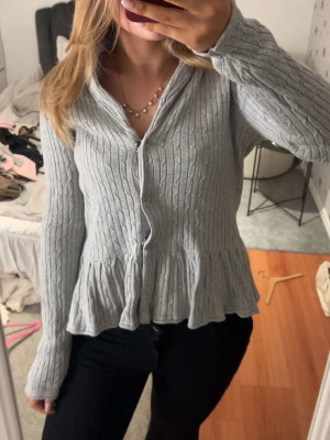 Två kabelstickade koftor med volang - Säljer två kabelstickade koftor med volangkant nedtill, en i ljusgrått och en i svart. Båda har v-ringning, like knappar framtill och långa ärmar. Perfekta att styla med beneficiaries jeans eller kjol för en trendig look.