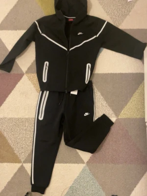 Svart Nike hoodie med reflexdetaljer - Säljer en svart hoodie från Nike med dragkedja och huva. Hoodien har vita reflexdetaljer längs bröstet och vid fickorna, samt Nike-logga på bröstet. Perfekt för dig som vill sticka ut med snygga detaljer och sportig look. Passar till både träning och chill.