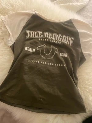 True Religion t-shirt med tryck - Riktig t-shirt från True Religion med kontrastfärgade ärmar och stort tryck på bröstet. T-shirten är olivgrön med vita ärmar och har en klassisk passform. Har inte använts mycket💕köpes för 300kr