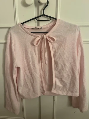 Ljusrosa kofta med rosett - Supersöt ljusrosa kofta med lång ärm och knytband i form av en rosett framtill. Tillverkad i ett mjukt, fluffigt material som känns riktigt cozy. Perfekt att slänga över en topp för en gullig och avslappnad look.