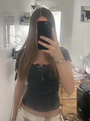 Svart ribbad topp med knappar - En svart ribbad topp med korta ärmar och knappdetaljer framtill. Snygg och enkel design som är lätt att matcha. Materialet är mjukt och stretchigt, perfekt för en avslappnad look.