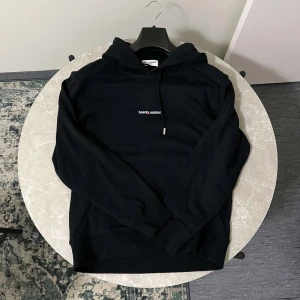 Svart hoodie från Saint Laurent - Snygg svart hoodie från Saint Laurent med logotyp på bröstet. Den har en klassisk design med dragsko i huvan och ribbade muddar vid ärmslut och nederkant. Perfekt för en stilren look. Hoodien är i bra skick. 