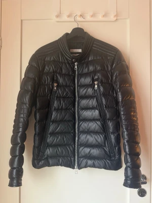 Svart skinn/dun jacka  från Moncler - Säljer en svart puffer dun jacka från Moncler i skinn med coola quiltade detaljer och två snedställda dragkedjefickor framtill. Jackan har hög krage och långa ärmar med knappdetaljer vid ärmslut. Perfekt för dig som vill ha en stilren och exklusiv look. Lite små skador på kanten av ena armen men inget som syns 