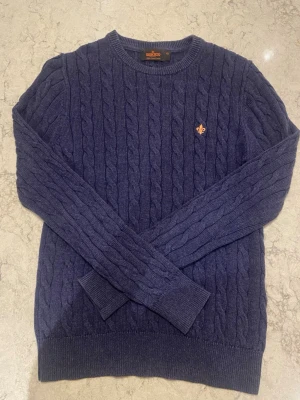 Blå kabelstickad tröja från Morris - Snygg blå kabelstickad tröja från Morris Stockholm i 100% pima bomull. Tröjan har rund halsringning, ribbade muddar och en liten orange logga broderad på bröstet. Perfekt för dig som gillar klassisk och stilren design.