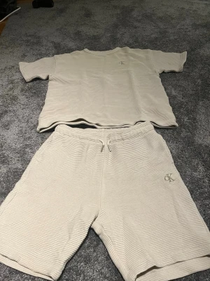 Beige ribbat set från Calvin Klein - Snyggt ribbat set från Calvin Klein i beige. Setet består av en t-shirt med kort ärm och matchande shorts med dragsko i midjan. Båda delarna har diskret CK-logga broderad framtill. Materialet är mjukt och stretchigt för maximal komfort.