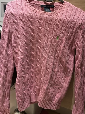 Rosa kabelstickad tröja Ralph Lauren - Kabelstickad rosa tröja från Ralph Lauren med rund halsringning och grönt broderat logotyp på bröstet. Tröjan har långa ärmar och ribbade muddar vid ärmslut och nederkant. Perfekt för dig som gillar klassisk och stilren look.