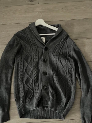 Mörkgrå kabelstickad kofta - Mysig mörkgrå kofta med kabelstickat mönster och stora svarta knappar framtill. Koftan har sjalkrage och ribbade muddar vid ärmslut och nederkant. Perfekt för lager på lager och en stilren look. Skriv för fler bilder 