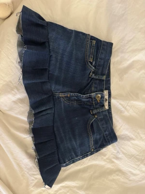 Minikjol fornarina - Säljer en mörkblå jeanskjol från fornarina med volangkant nedtill. Kjolen har klassiska fickor fram och bak, synliga sömmar och knappgylf. Perfekt för dig som gillar en edgy och trendig stil.